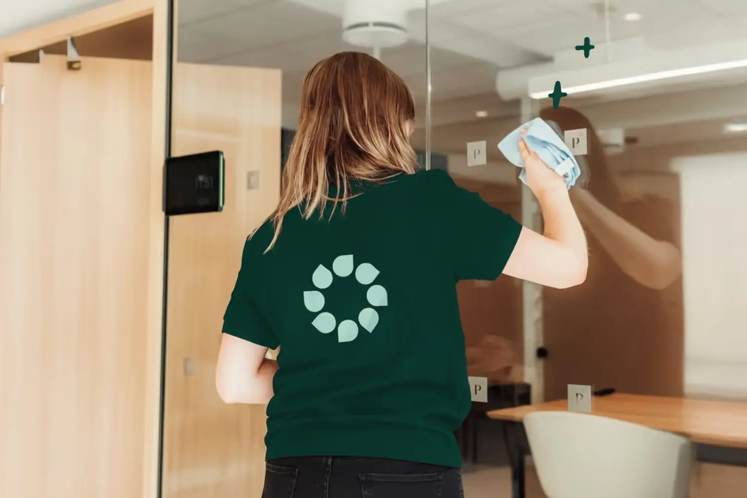 Person med grønn t-skjorte vasker glassvegg inne i et lyst moderne kontorlandskap.